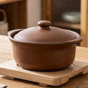 Può includere: Una pentola da cucina in ceramica marrone con coperchio, appoggiata su un sottopentola quadrato in legno. La pentola è su un tavolo di legno, con bacchette, tazze da tè e un tovagliolo di lino piegato. La scena suggerisce una cucina o una sala da pranzo.