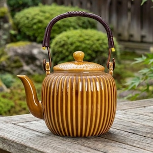 Puede incluir: Una tetera de cerámica marrón con detalles acanalados verticales y una tapa a juego. La tetera tiene un asa trenzada marrón oscuro y un pico curvo. Está sobre una mesa de madera en un jardín.