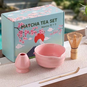 May include: Matcha tea set with a pink bowl, whisk, and holder, plus a bamboo scoop. The box is light blue with cherry blossom illustrations and the text "Matcha Tea Set". The set is displayed on a white and gray patterned surface.