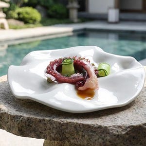 以下が含まれることがあります： 白い有機的な形の皿には、料理が盛り付けられています。料理は、濃い赤色のタコの触手、キュウリのスライス、少量のソースで構成されています。皿は石の表面に置かれ、背景にはプールと日本庭園があります。