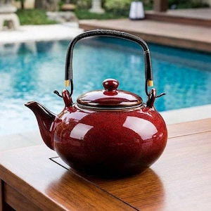 Könnte beinhalten: Eine glänzende, kastanienbraune Keramik-Teekanne mit schwarzem Griff und goldenen Akzenten. Die Teekanne hat einen abgerundeten Körper, einen Ausguss und einen Deckel mit einem kleinen Knopf. Sie steht auf einer Holzoberfläche, mit Pool und Garten im Hintergrund.