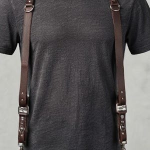 May include: A dark brown leather dual camera harness worn over a charcoal gray short-sleeved t-shirt. The harness features adjustable straps, metal buckles, and silver-toned hardware, with two black digital cameras attached to the lower clips.