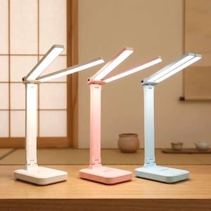 May include: Three modern LED desk lamps in white, pink, and light blue. Each lamp has a rectangular base, a vertical stem, and a pivoting light panel. The lamps are on a wooden surface.