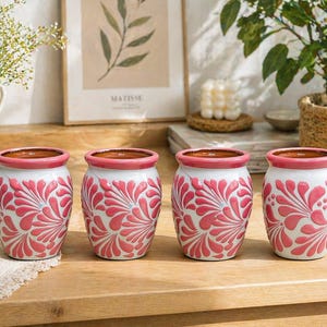 May include: Four white ceramic pots with a pink floral design. The pots have a pink rim and interior. The pots are arranged in a row on a wooden surface. The pots are suitable for indoor plants.