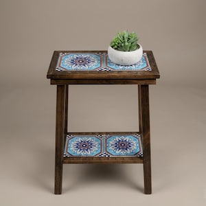 May include: A small, two-tiered wooden side table with a dark brown finish. The top and lower shelf feature blue and white patterned tiles. A small, white ceramic pot with a green succulent plant sits on the top shelf. The table is 24 inches tall.