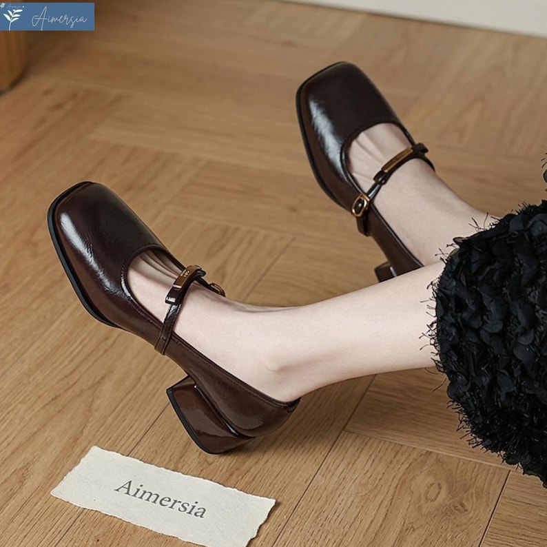 May include: A pair of dark brown leather Mary Jane style shoes with square toes and block heels. Each shoe has a strap with a gold-colored buckle. The shoes are on a wooden floor. The brand name "Aimersia" is visible on a small tag.