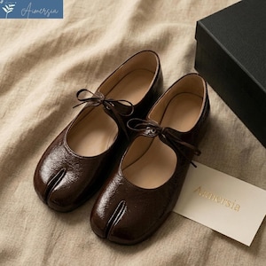 Puede incluir: Un par de bailarinas Mary Jane de cuero marrón con puntera dividida y lazos. Los zapatos tienen un acabado brillante y una puntera redondeada. El nombre de la marca "Aimersia" es visible en una tarjeta pequeña.