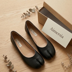 May include: A pair of black split-toe ballet flats with a low heel, displayed on a wooden surface. The shoes are next to an open cardboard box and a card that reads "Aimersia". The shoes have the brand name "OKLIBO" printed on the insole.