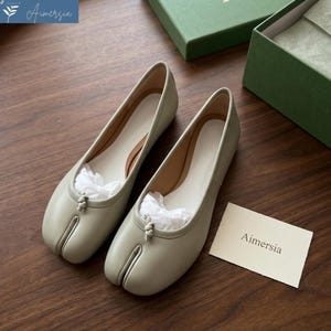 May include: A pair of light green leather ballet flats with a split toe design. The shoes have a rounded toe and a small knot detail at the top. The interior is a light cream color. The brand name "Aimersia" is visible on a small card.