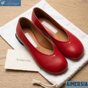 K&ouml;nnte beinhalten: Ein Paar rote Leder-Ballerinas mit runder Spitze, die auf einem wei&szlig;en Stoffbeutel liegen. Die Schuhe haben ein kleines wei&szlig;es Etikett auf der Oberseite und ein kleines wei&szlig;es Nahtdetail. Der Markenname "Aimersia" ist sichtbar.