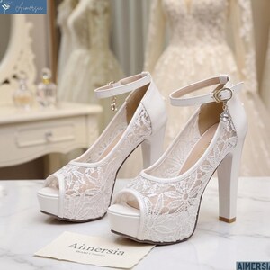 May include: White peep-toe platform heels with a delicate floral lace overlay. The heels have a thick block heel and an ankle strap with a small charm. The brand name "Aimersia" is visible on a small card.
