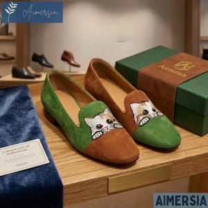 May include: A pair of suede loafers with a cat face embroidered on the toe. The shoes are brown and green, with a green vamp and a brown toe. The shoes are displayed on a wooden shelf with a box and a certificate.
