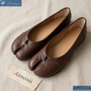 Può includere: Un paio di ballerine in pelle marrone con punta divisa. Le scarpe hanno un design a punta arrotondata con una divisione tra l'alluce e le altre dita. Il nome del marchio "Aimersia" è visibile.