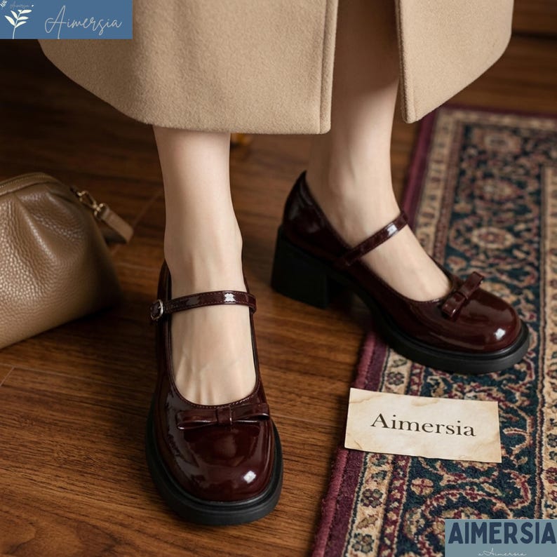 May include: Burgundy Mary Jane style shoes with a bow detail and a block heel. The shoes have a glossy finish and a strap that buckles across the top of the foot. The shoes are on a patterned rug. The brand name "Aimersia" is visible.