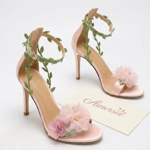 Könnte beinhalten: Hellrosa High-Heel-Sandalen mit Blumen- und Blattdetails. Die Sandalen haben einen Knöchelriemen, der mit grünen Blättern und einer Schnalle verziert ist. Der Zehenbereich ist mit rosa Stoffblumen verziert. Die Absätze sind etwa 10 cm hoch.