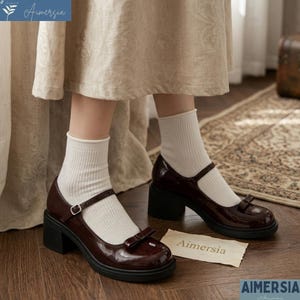 May include: Burgundy Mary Jane style shoes with a bow detail and a block heel. The shoes have a glossy finish and a strap that buckles across the top of the foot. The shoes are paired with white socks.