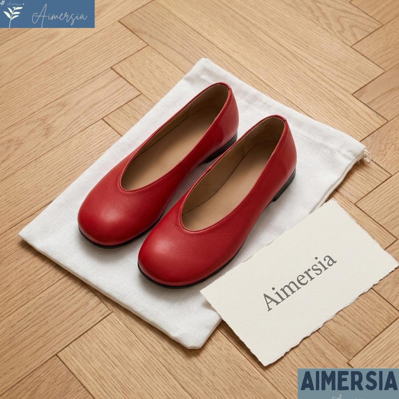 K&ouml;nnte beinhalten: Ein Paar rote Leder-Ballerinas mit runder Spitze, auf einem wei&szlig;en Stoffbeutel. Die Schuhe haben eine glatte, gl&auml;nzende Oberfl&auml;che. Eine Karte mit dem Wort "Aimersia" befindet sich rechts.