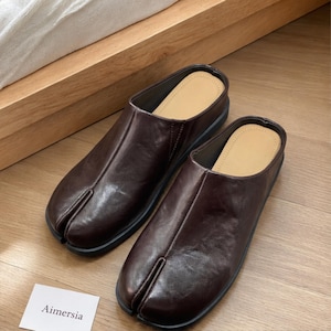 May include: A pair of brown leather slip-on shoes with a split-toe design. The shoes have a black sole and a tan insole. The shoes are sitting on a wooden surface. The brand name "Aimersia" is on a white card.