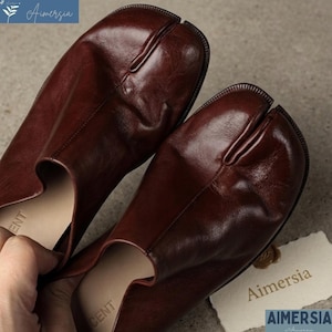 Pode incluir: Um par de sapatos de couro castanho escuro com biqueira dividida. Os sapatos têm um design único com uma divisão entre o dedão do pé e os outros dedos. Os sapatos são feitos de couro liso e brilhante.