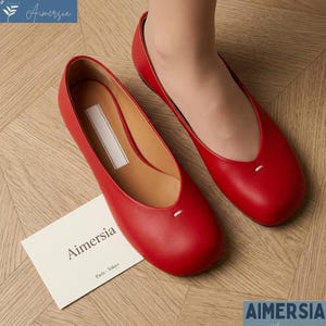 K&ouml;nnte beinhalten: Ein Paar leuchtend rote Leder-Ballerinas mit runder Spitze und einem kleinen wei&szlig;en Nahtdetail. Die Schuhe werden auf einer hellen Holzoberfl&auml;che pr&auml;sentiert, mit einer Karte mit der Aufschrift "Aimersia Paris-Tokyo".