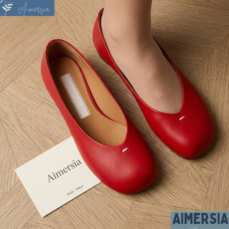 K&ouml;nnte beinhalten: Ein Paar leuchtend rote Leder-Ballerinas mit runder Spitze und einem kleinen wei&szlig;en Nahtdetail. Die Schuhe werden auf einer hellen Holzoberfl&auml;che pr&auml;sentiert, mit einer Karte mit der Aufschrift "Aimersia Paris-Tokyo".