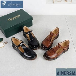 Könnte beinhalten: Zwei Paar schwarze und zwei Paar braune Mary Jane-Schuhe aus Leder mit T-Riemen-Design. Die Schuhe haben einen Schnallenverschluss und Fransen. Die Schuhe werden mit einer grünen Schachtel mit dem Text "Aimersia" präsentiert.