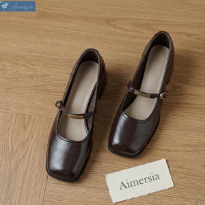 May include: Pair of dark brown leather Mary Jane style shoes with square toes and small heels. Each shoe has a strap across the top with a gold-colored buckle. The shoes are on a wooden surface. A small card with the word "Aimersia" is on the surface.