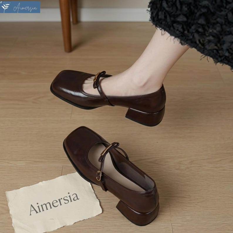 May include: Brown leather Mary Jane style shoes with a square toe and small block heel. The shoes have a strap across the top with a gold-colored buckle. The word "Aimersia" is printed on a piece of paper.