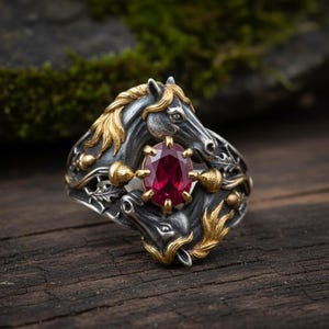 Puede incluir: Anillo de plata con diseño de dos cabezas de caballo, detalles dorados y un gran rubí ovalado. El anillo presenta detalles intrincados y una estética vintage, ideal para entusiastas de la equitación o amantes de la joyería única.
