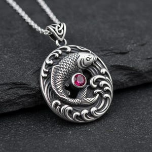 Könnte beinhalten: Silberne Halskette mit Anhänger im Koi-Karpfen-Design. Der runde Anhänger zeigt einen Fisch, der durch Wellen schwimmt, mit einem roten Edelstein in der Mitte. Die Halskette ist ebenfalls aus Silber.