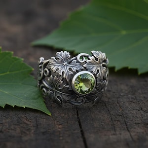 Könnte beinhalten: Ein silberner Ring mit Blättermuster und einem runden, hellgrünen Edelstein. Der Ring liegt auf einer dunklen Holzoberfläche mit grünen Blättern im Hintergrund. Das filigrane Blättermuster des Rings umschließt das Band.