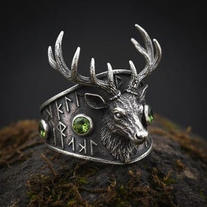May include: A silver-toned ring featuring a detailed deer head with large antlers. The ring has green gemstones and engraved text around the band. The ring is set on a dark, textured surface.
