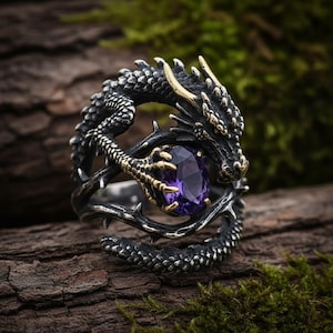 Könnte beinhalten: Ein detaillierter silberner Drachenring mit goldenen Akzenten und einem großen, ovalen, lila Edelstein. Der Drache umschlingt den Finger, seine strukturierten Schuppen und Krallen halten den Edelstein. Der Ring ist vor einem natürlichen Holzgrund platziert.