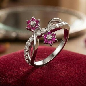 Puede incluir: Un anillo plateado con un diseño floral. El anillo presenta dos grupos de gemas rosas en forma de flor, cada uno rodeado de piedras transparentes más pequeñas. La banda está adornada con piedras transparentes. El anillo está sobre una superficie de terciopelo rojo.