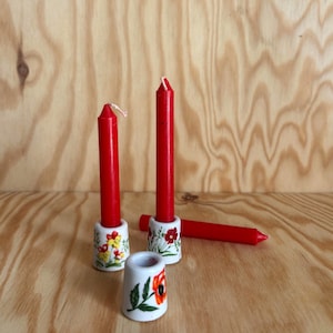 Peut inclure: Bougies rouges effilées avec des porte-bougies en céramique florale. Les supports sont blancs avec des motifs floraux colorés. Une bougie est posée horizontalement. Le fond est un grain de bois clair.