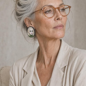 May include: A person wearing brown-framed spectacles and a beige blazer. The person has grey hair in a bun and is wearing oval earrings with a portrait design. The earrings feature a blonde woman with a green collar.