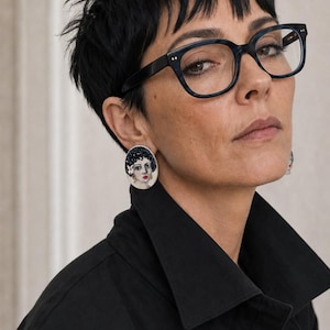 May include: A person wearing black-framed spectacles and a black collared shirt. They are also wearing large, oval earrings with a portrait design. The earrings feature a woman's face with dark hair and red lipstick. The person has short, dark hair.