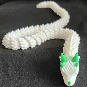 May include: A white, articulated dragon figurine with a textured, scale-like body. The dragon's head features green horns and accents. The figurine is made of a 3D-printed material and is posed on a dark surface.