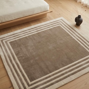 May include: A rectangular area rug featuring a solid taupe center panel framed by concentric borders in alternating shades of cream and taupe. The rug has a flat, uniform texture and clean, straight edges, creating a geometric pattern on the floor.
