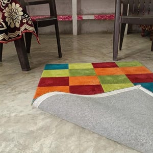 May include: A rectangular area rug featuring a multi-coloured grid pattern of squares in shades of red, orange, lime green, and teal. One corner of the rug is folded over to reveal a light grey, textured fabric backing with a white stitched border edge.