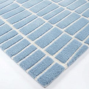 May include: A light blue rectangular rug with a geometric pattern. The rug features a repeating design of light blue rectangles separated by off-white lines. The rug has a soft, textured appearance and is set against a white background.