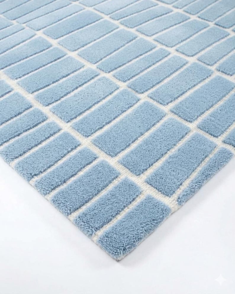 May include: A light blue rectangular rug with a geometric pattern. The rug features a repeating design of light blue rectangles separated by off-white lines. The rug has a soft, textured appearance and is set against a white background.