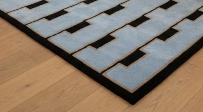 K&ouml;nnte beinhalten: Ein rechteckiger Teppich mit einem sich wiederholenden geometrischen Muster aus hellblauen, ineinandergreifenden Formen, die von d&uuml;nnen beigen R&auml;ndern auf einem einfarbigen schwarzen Hintergrund umrandet sind und ein strukturiertes Design bilden.
