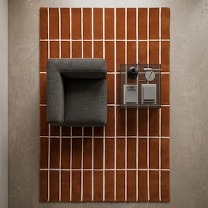 May include: A rectangular area rug featuring a burnt orange background with a white grid pattern of vertical and horizontal lines. The rug is positioned under a dark gray armchair and a square glass side table with books and a small vase.