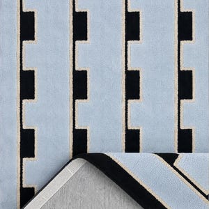 K&ouml;nnte beinhalten: Ein rechteckiger Teppich mit einem sich wiederholenden Muster aus hellblauen vertikalen Streifen, die durch schwarze rechteckige Bl&ouml;cke und d&uuml;nne cremefarbene Umrandungen getrennt sind. Eine Ecke ist umgeschlagen und zeigt die hellgraue R&uuml;ckseite.