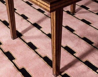 May include: A wooden table with a polished, light brown burl wood top and dark brown edges. The table features four straight, vertically grooved legs made of matching light and dark brown wood, standing on a pink rug with black geometric patterns.