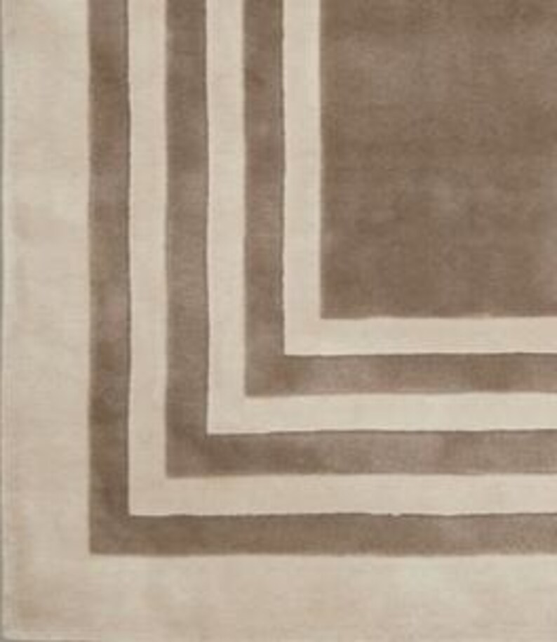 May include: A rectangular area rug featuring a geometric pattern of alternating taupe and cream-colored bands. The design consists of concentric right-angled stripes that frame a solid taupe center, creating a layered border effect across the surface.