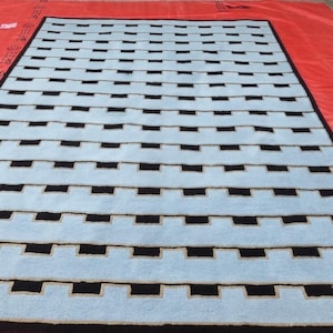 K&ouml;nnte beinhalten: Ein rechteckiger Teppich mit einem sich wiederholenden geometrischen Muster aus hellblauen horizontalen Streifen, die von schwarzen rechteckigen Bl&ouml;cken unterbrochen werden. Der Teppich ist von einem schwarzen Rand umgeben.