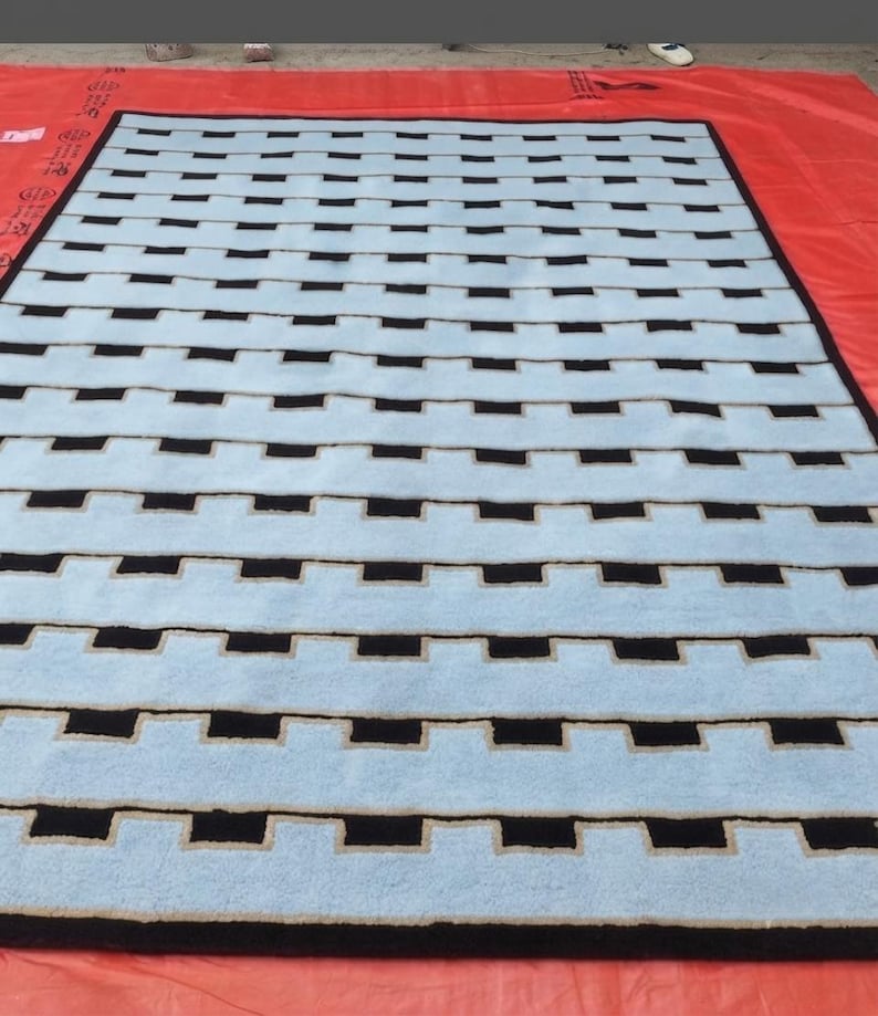 K&ouml;nnte beinhalten: Ein rechteckiger Teppich mit einem sich wiederholenden geometrischen Muster aus hellblauen horizontalen Streifen, die von schwarzen rechteckigen Bl&ouml;cken unterbrochen werden. Der Teppich ist von einem schwarzen Rand umgeben.