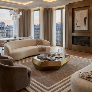 May include: A living room featuring a curved cream sofa, two brown armchairs, and a geometric brass coffee table with a dark stone top. The space includes a fireplace with a framed abstract painting, a crystal chandelier, and large windows.